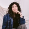 Mujer con cabello rizado, vistiendo una chaqueta vaquera y un reloj, sentada con la mano apoyada en la barbilla frente a un fondo de tela drapeado.